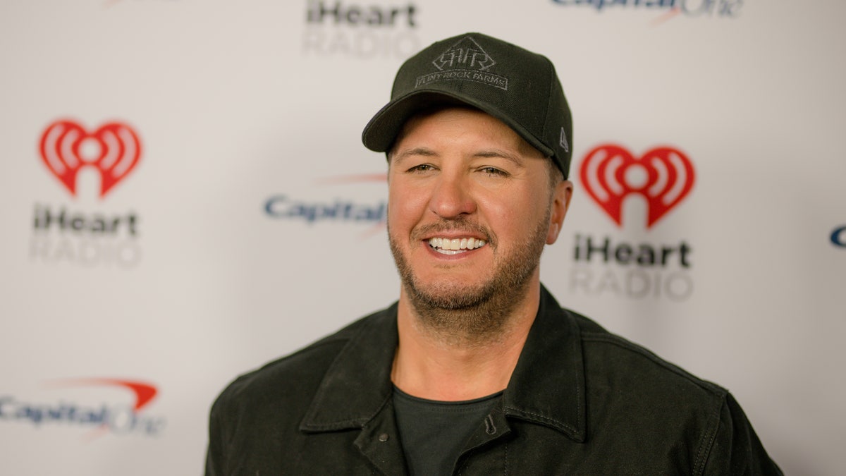 Luke Bryan in a black jean jacket and black hat on the carpet for iHeartCountry festival