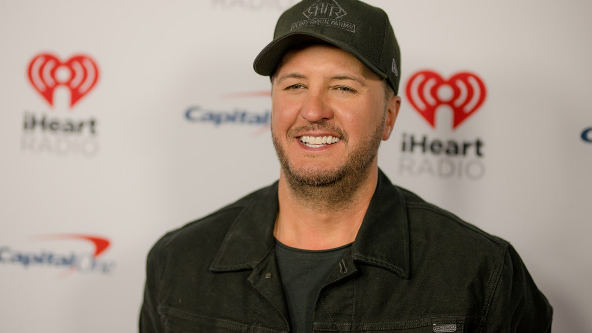 Luke Bryan in a black jean jacket and black hat on the carpet for iHeartCountry festival