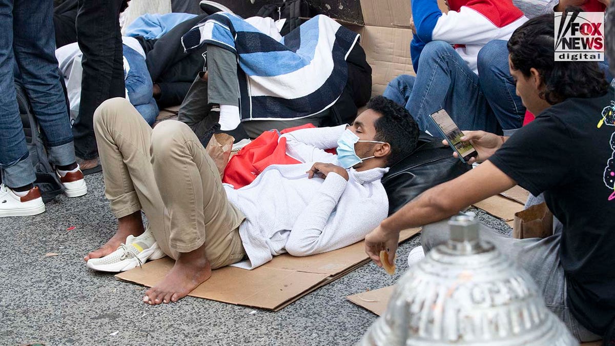 Migrants rest on cardboard panels on sidewalk outside hotel