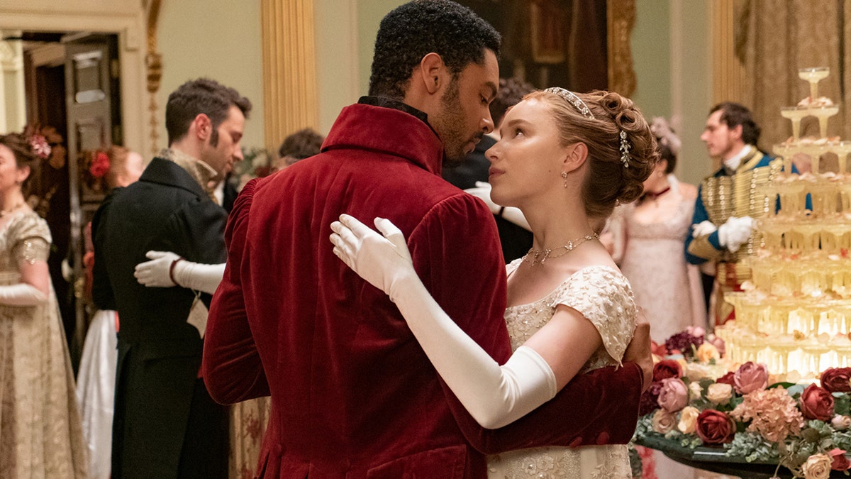 The Duke of Hastings wearing a burgundy coat dancing with Daphne Bridgerton wearing a lace white dress