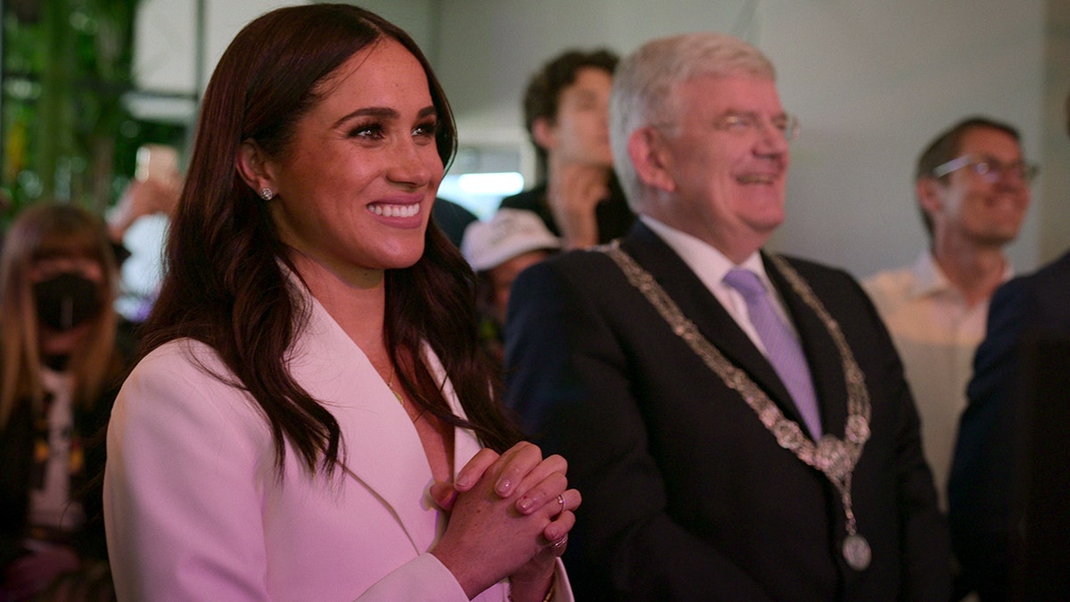 Meghan Markle wearing a white blazer standing next to a man wearing a suit and medals