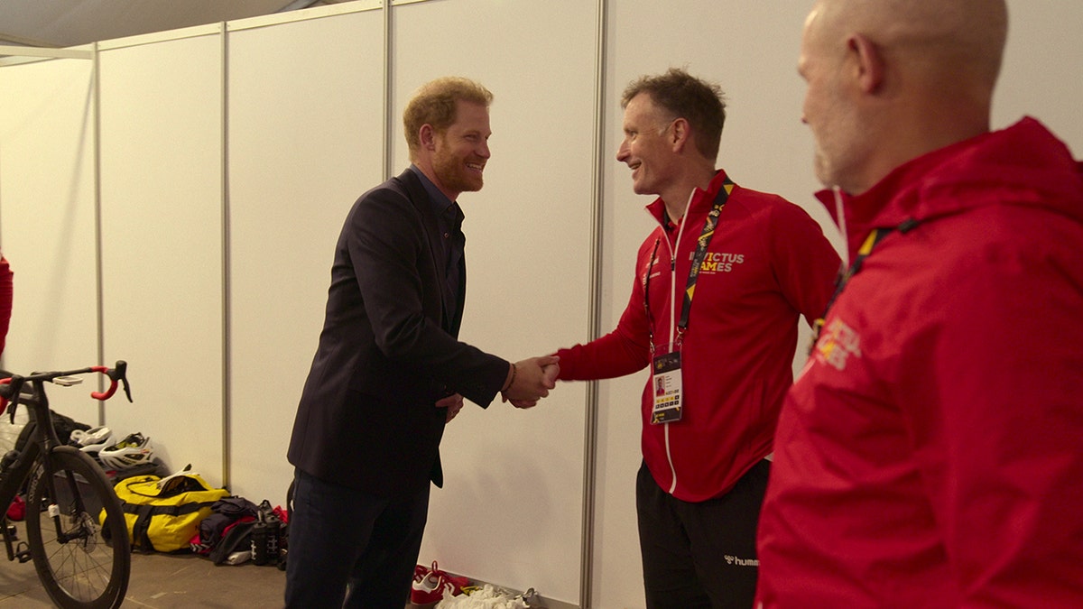 Prince Harry in a dark blazer shaking hands with a man in a red sweater