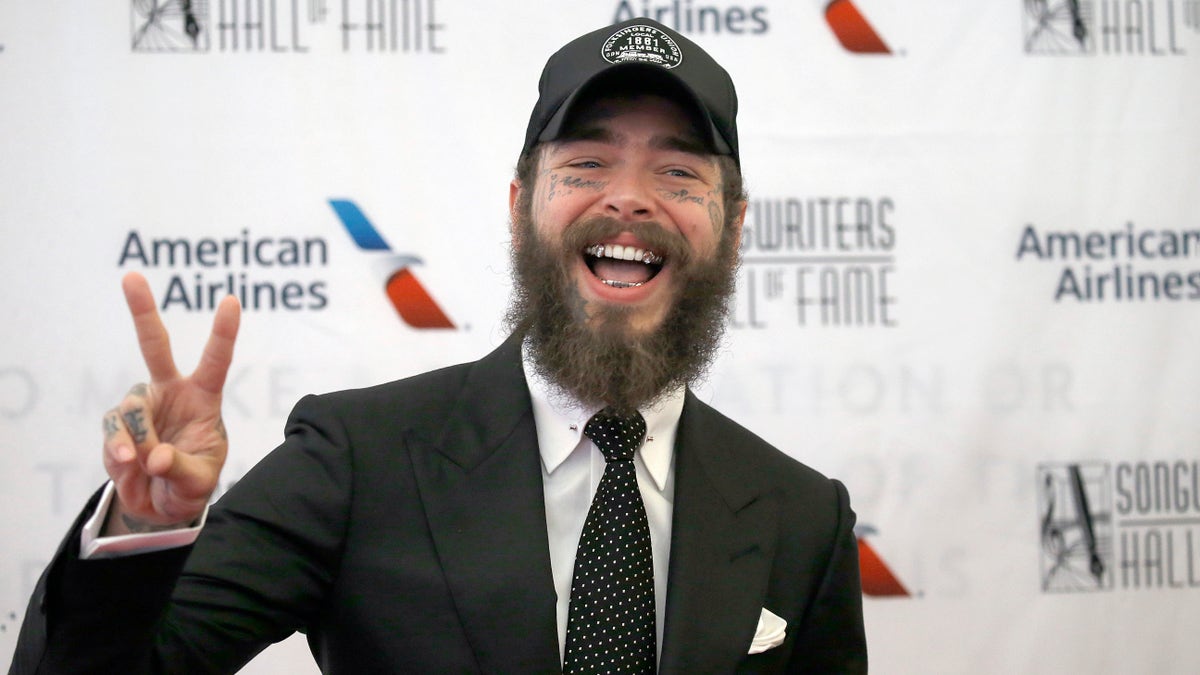 Post Malone makes a piece sign in a black suit and black baseball cap