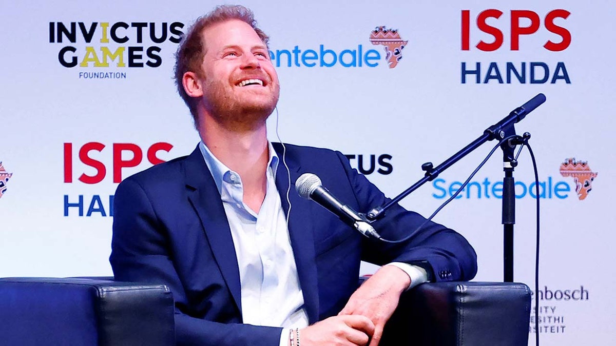 Prince Harry smiling wearing a navy blazer and a open button shirt