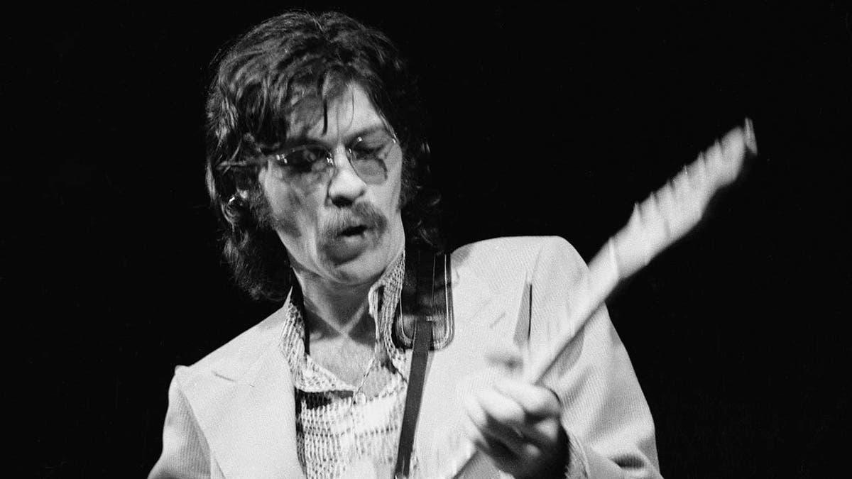 Robbie Robertson mengenakan setelan jas saat bermain gitar di atas panggung di London