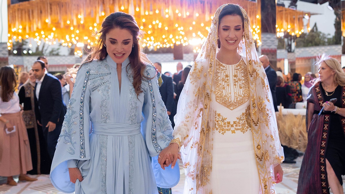 Queen Rania of Jordan wearing a light blue dress holding onto the hand of Princess Rajwa, who is wearing a white and gold dress