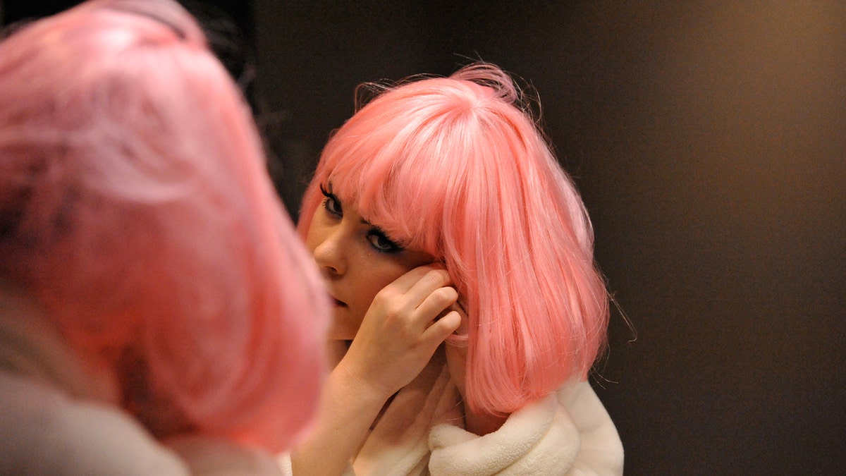 A close-up of Tasha Layton wearing a hot pink wig and a white robe applying eyeliner