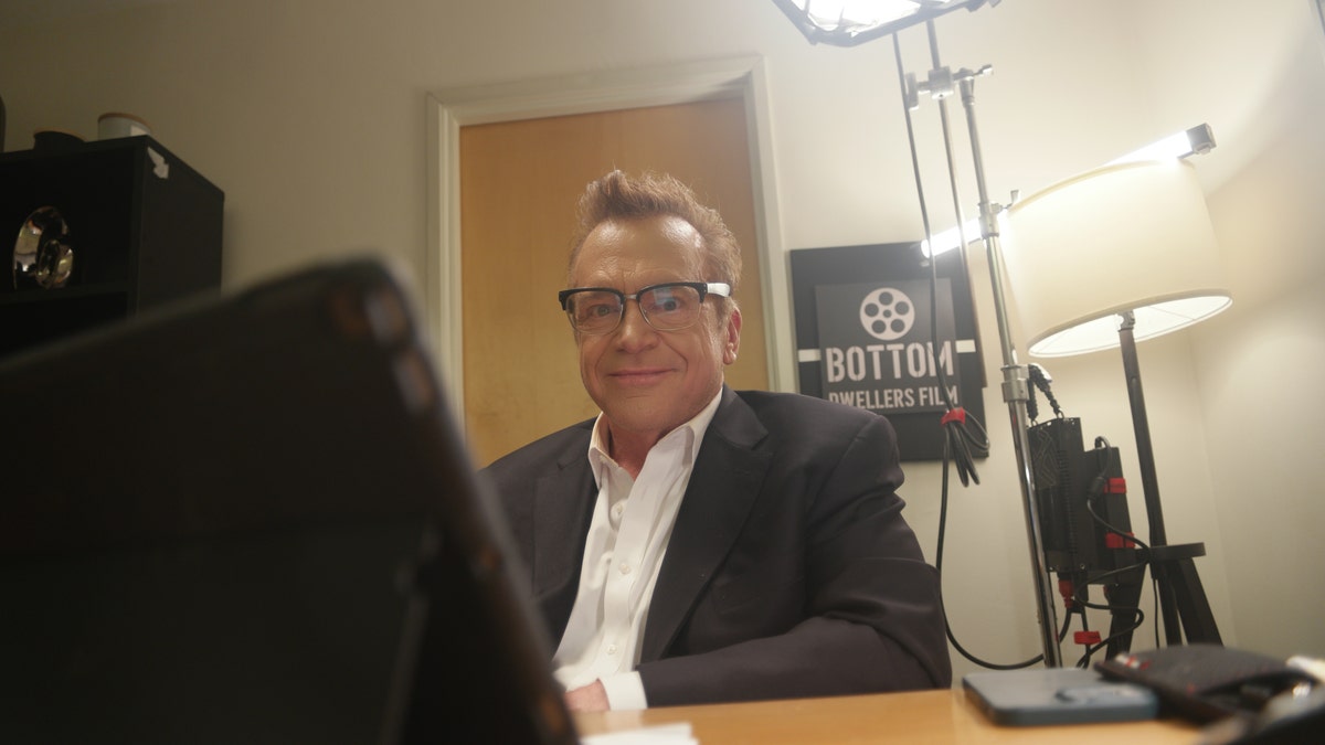 Tom Arnold sitting at a desk in character