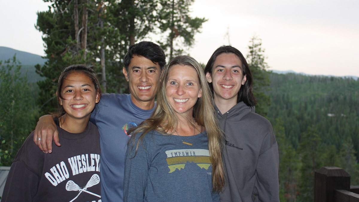 Wendi Lou Lee wearing a dark grey/blue sweater smiling next to her husband and two children in the woods