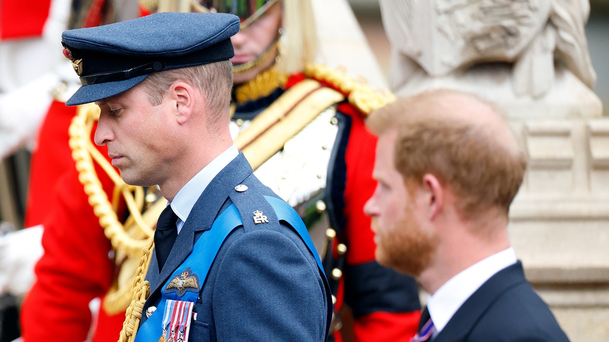 Prince William in a blue military suit looking away as Prince Harry looks at him in a morning suit