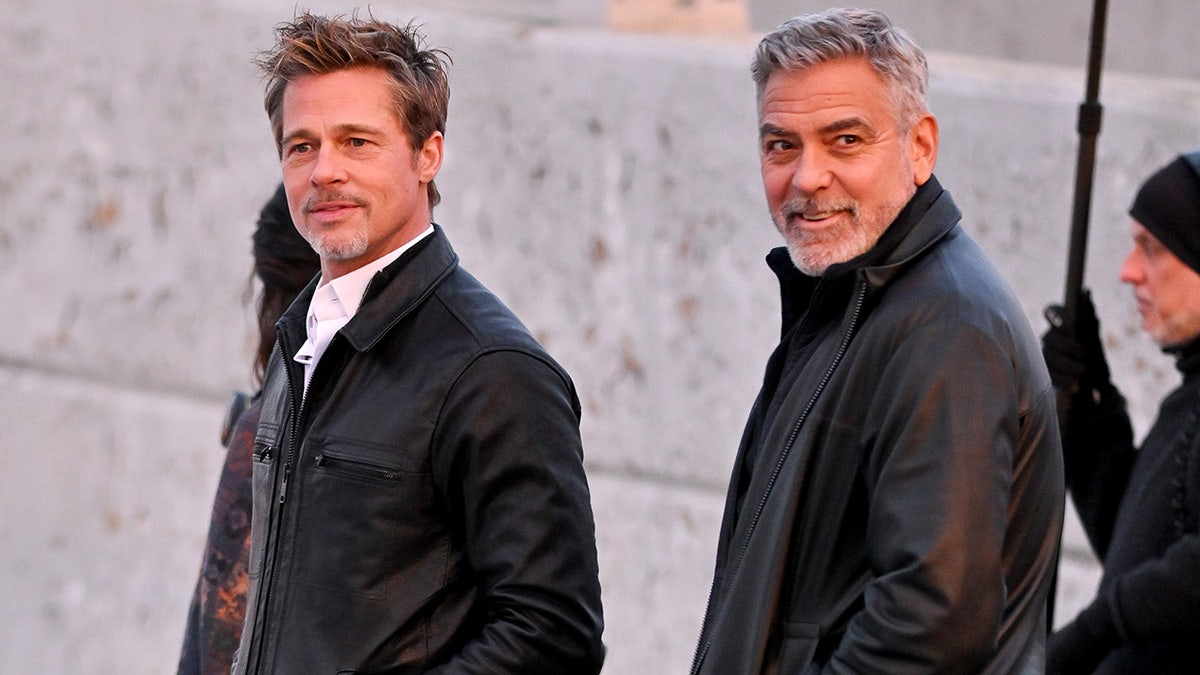 Brad Pitt in a white shirt and black jacket walks alongside George Clooney in a black jacket in New York on set of their movie "Wolves"