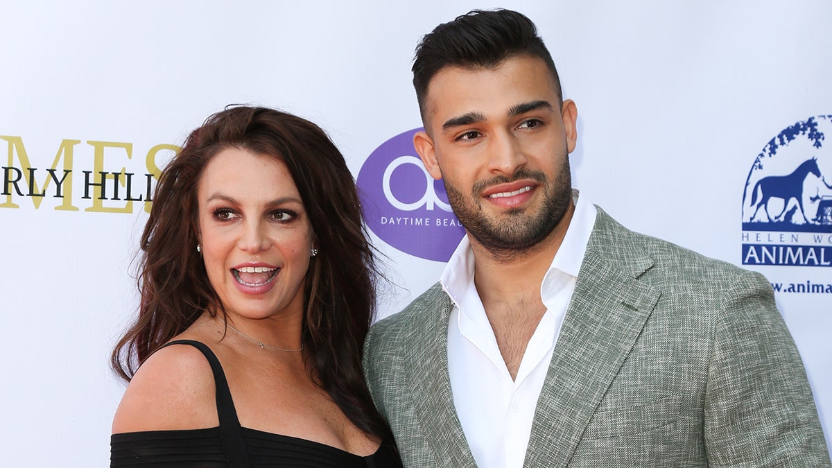 Britney Spears in a black dress looks to her right in a black dress as Sam Asghari looks to his left in a grey suit
