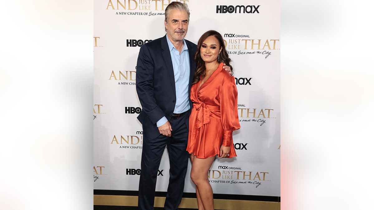 Chris Noth dengan kemeja biru dan setelan jas hitam di karpet bersama istrinya Tara Wilson dengan rok pendek oranye yang terbakar halus