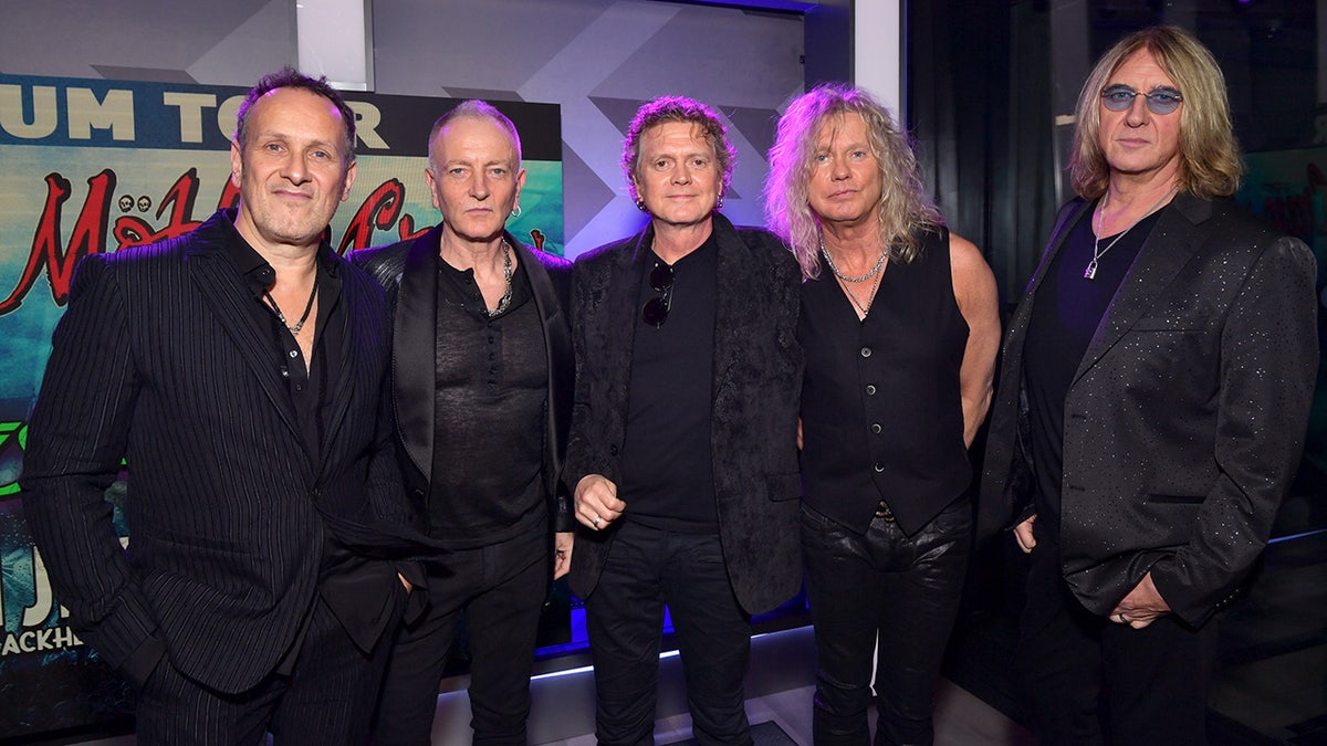 Vivian Campbell, Phil Collen, Rick Allen, Rick Savage, and Joe Elliott all in black outfits pose for a photo at a press conference announcing their joint tour with Mötley Crüe