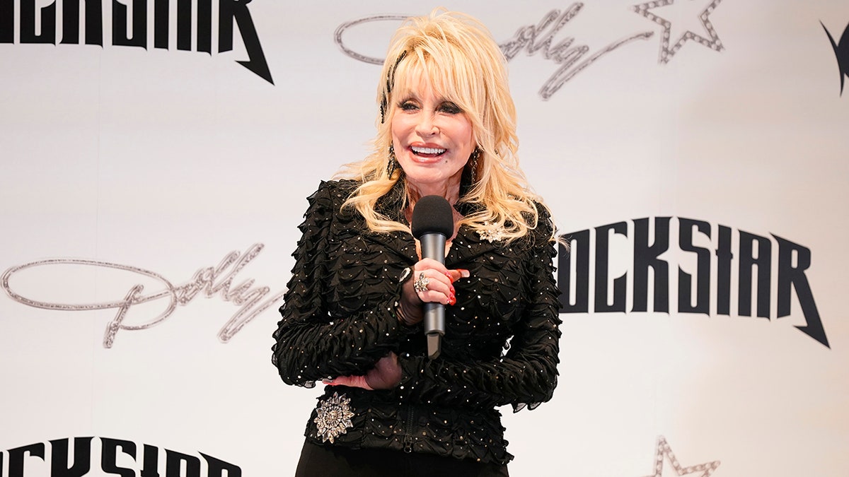 Dolly Parton in a black sparkle blazer and pantsuit speaks into the microphone