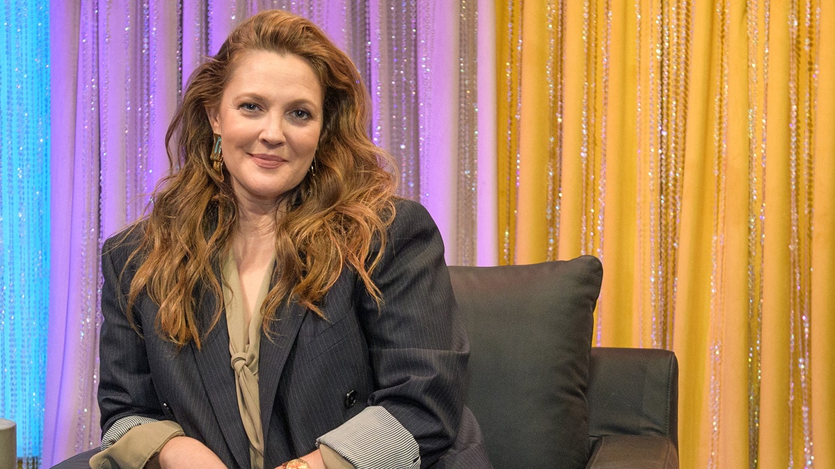 Drew Barrymore in a dark grey blazer with rolled up sleeves sits on a chair and soft smiles for the camera