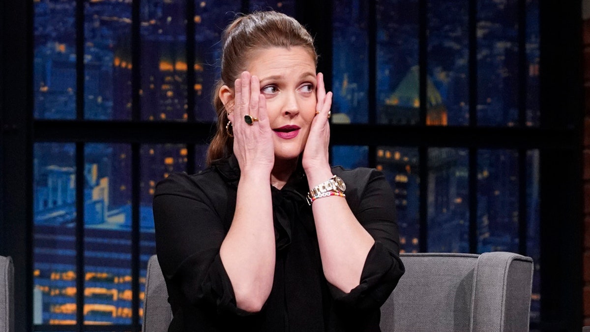 Drew Barrymore em um vestido preto coloca as mãos no rosto em perigo "Tarde da noite com Seth Meyers"