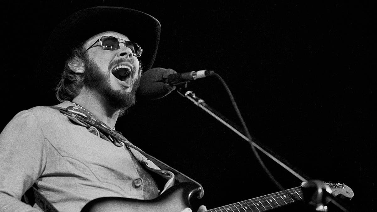 Black and white photo of Hank Williams Jr. passionately singing into the microphone wearing a black hat and sunglasses on stage