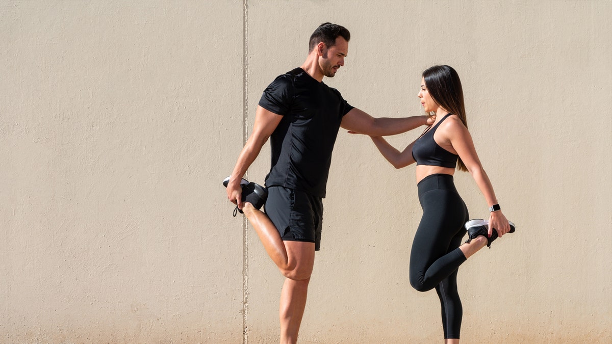 A tall man and short woman in gym attire stretch together.
