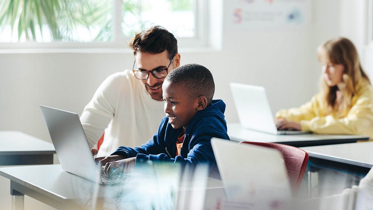 Male teacher helps a young boy with computer-based learning in a classroom setting.