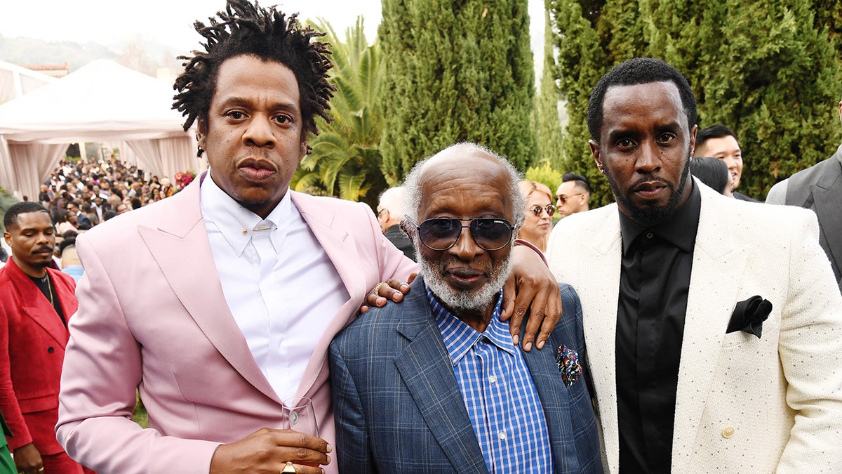 Jay-Z in a pink suit puts his arm around Clarence Avant and Diddy puts in a white suit and black shirt puts his arm around Clarence on the other side
