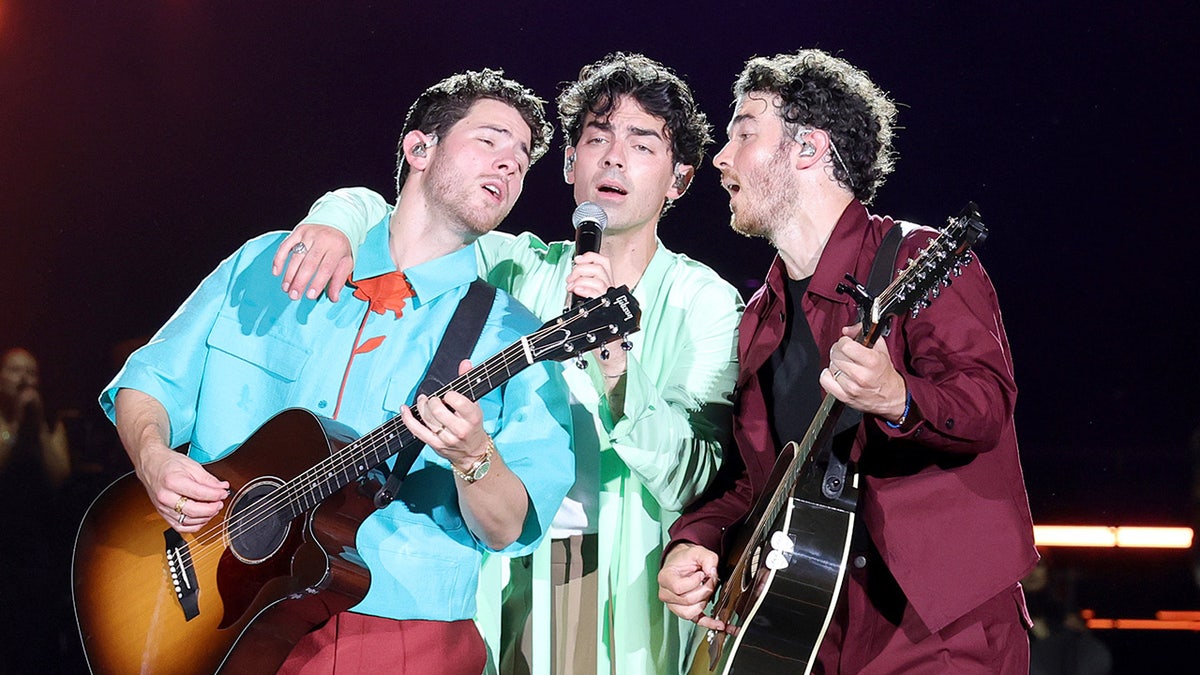 Nick Jonas playing the guitar leans back into Joe Jonas singing with Kevin Jonas, also playing the guitar leaning in to sing while on stage