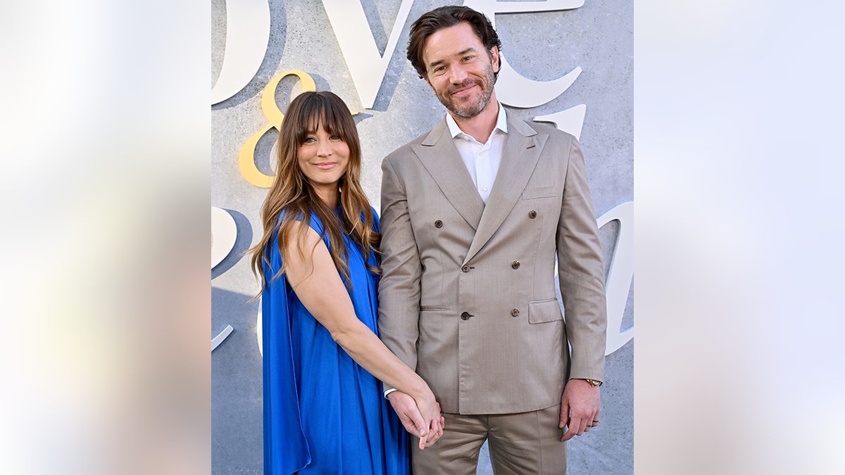 Kaley Cuoco in a long sleeveless blue silk dress holds hands with Tom Pelphrey on the carpet