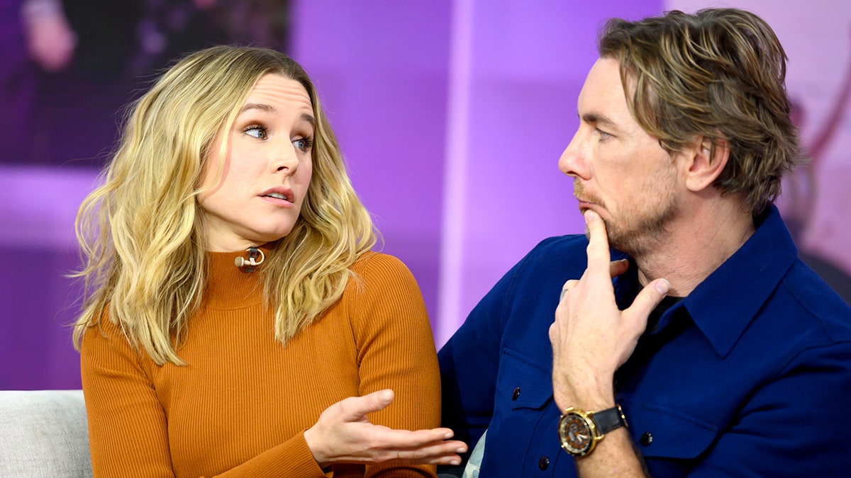 Kristen Bell in a burnt orange sweater looks towards Dax Shepard with a hand to his mouth while sitting on a couch during an interview
