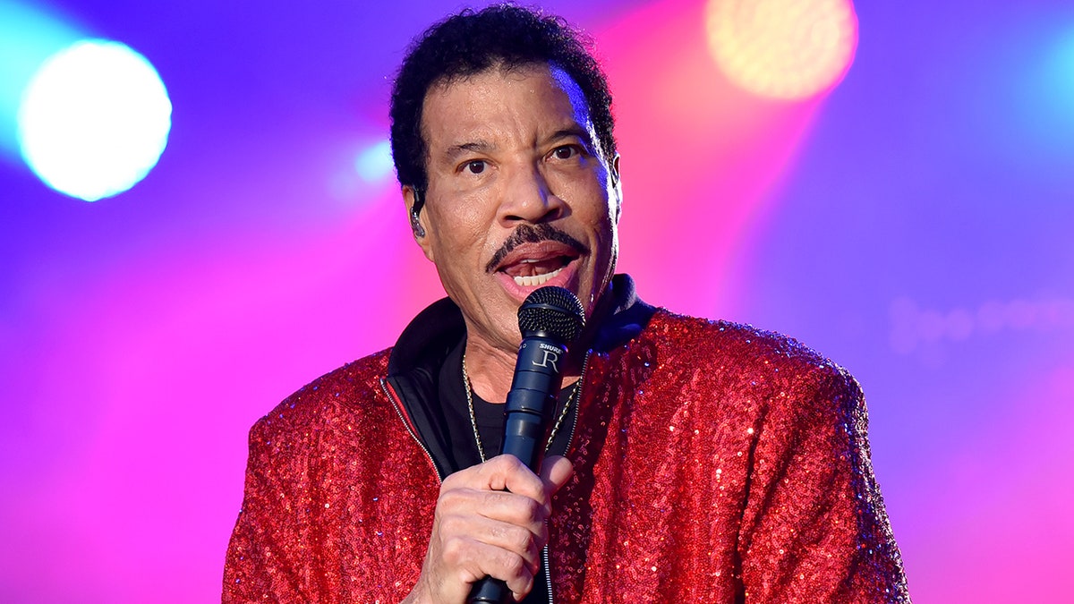 Lionel Richie speaks into the microphone on stage wearing a sparkly red jacket