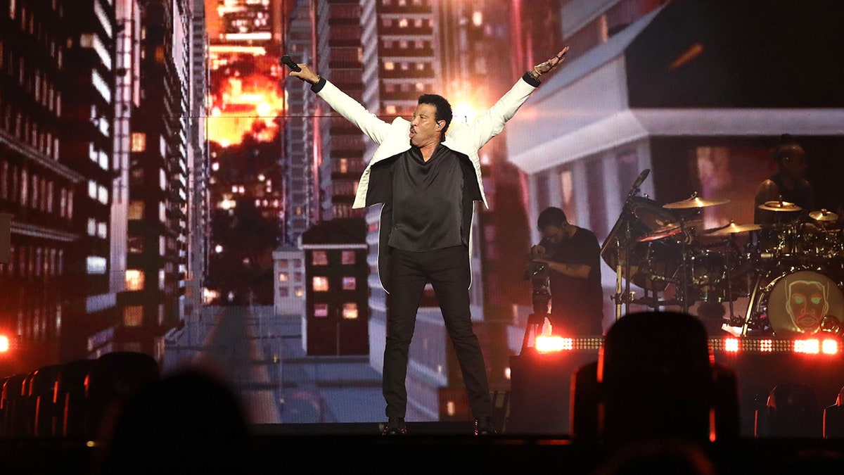 Lionel Richie wears a white jacket and black pants and shirt and puts his arms in the air on his tour with Earth, Wind & Fire