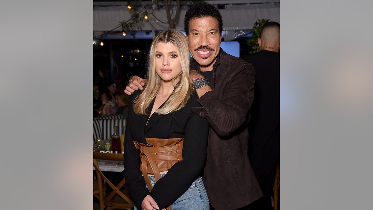 Sofia Richie in a black shirt and brown leather waist top soft smiles as her father Lionel Richie leans against her