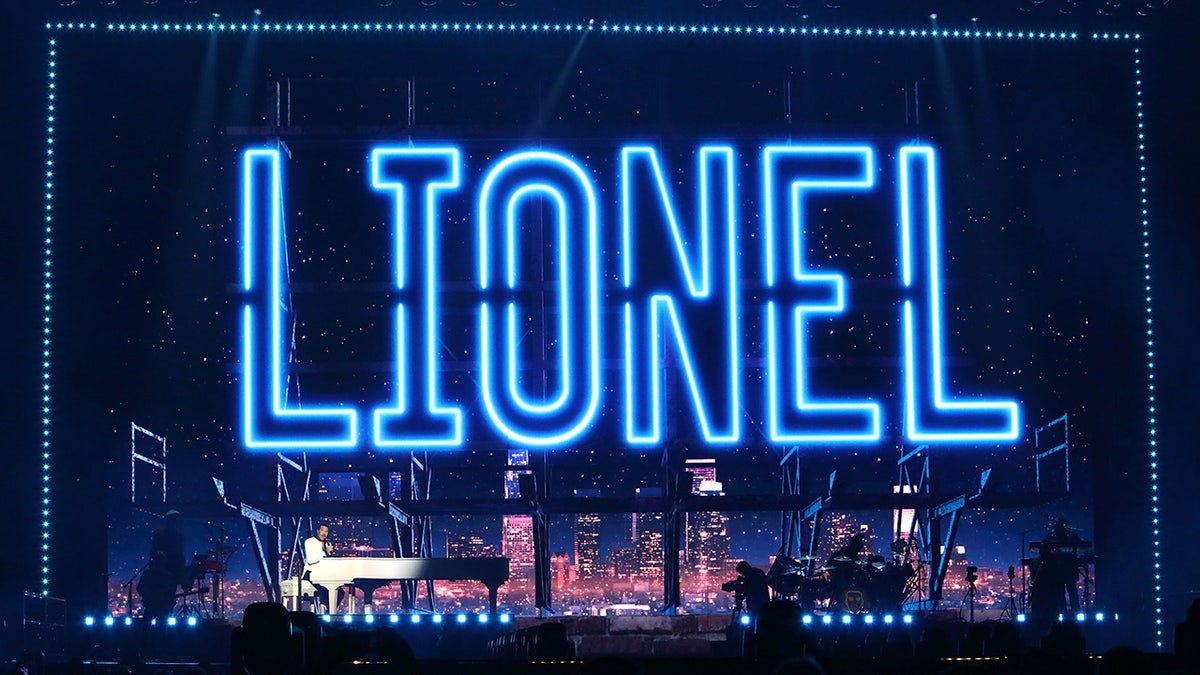 Lionel Richie on stage playing from a white piano with his name outlined in blue lights on the screen behind himawan