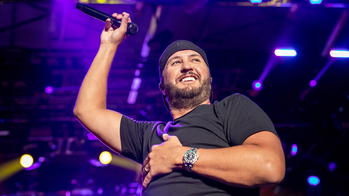 Luke Bryan in a black shirt and black backward hat raises his arm in the air over his head while on stage in Illinois
