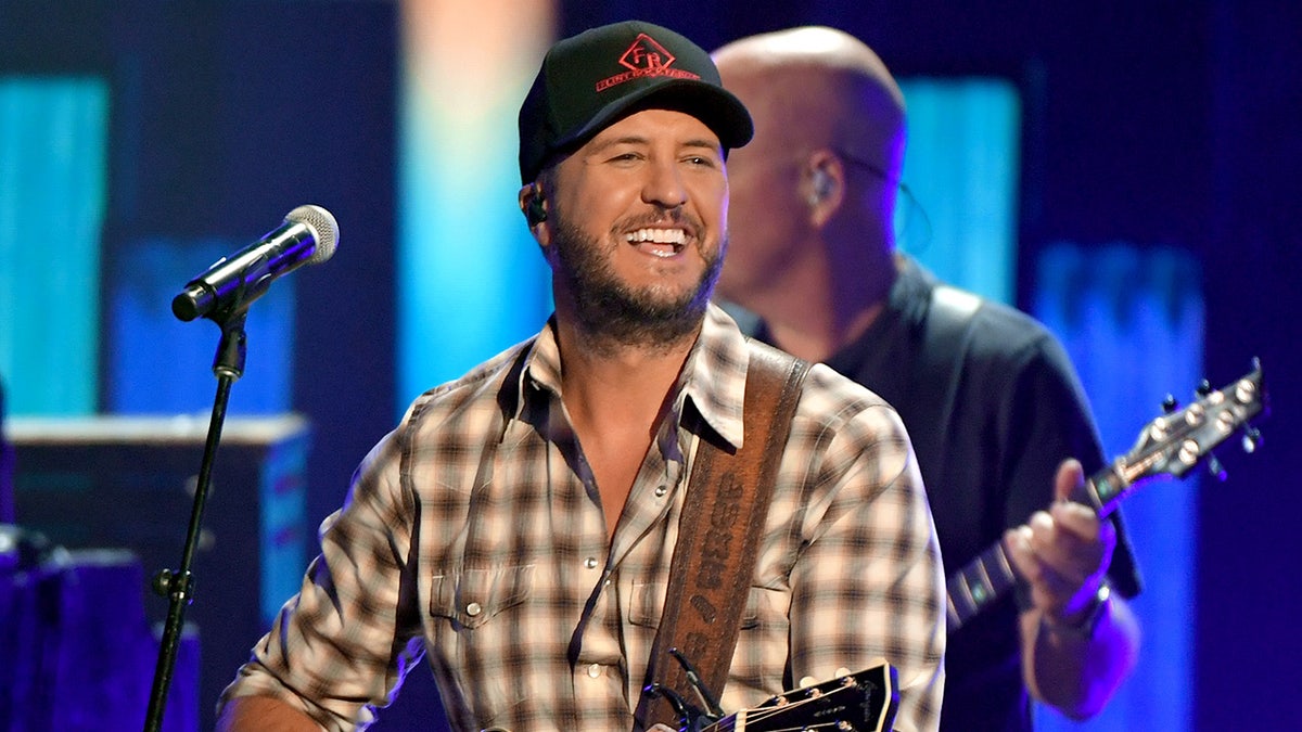 Luke Bryan in a checkered shirt and black cap plays the guitar at the ACM Awards