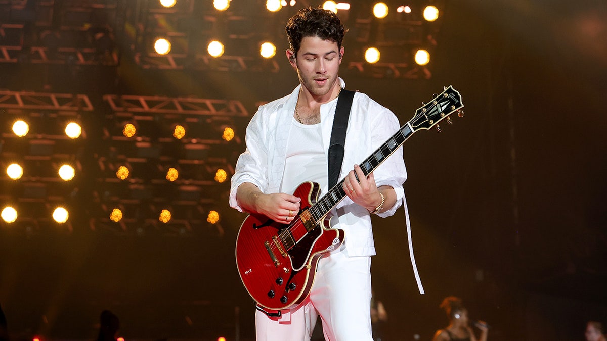 Nick Jonas in all white plays the guitar on stage and looks down at Yankee Stadium
