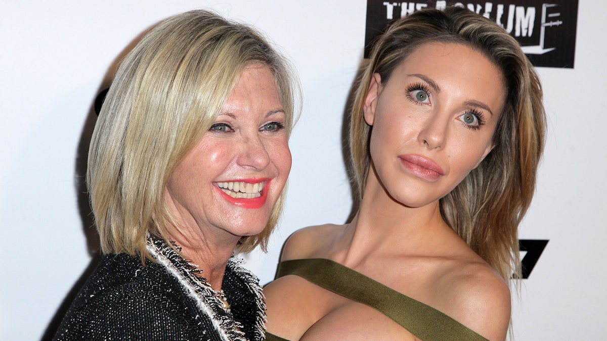 Olivia Newton-John smiles in a black blazer with her daughter Chloe in a green tube top with a large cutout at the top