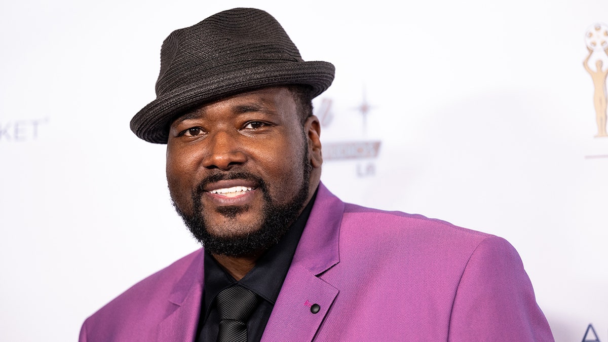 Quinton Aaron in a purple suit and black hat that is slightly tiled on the carpet
