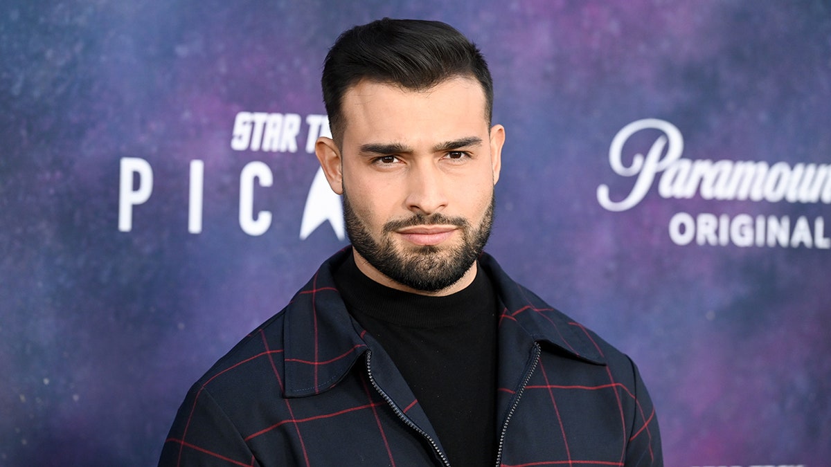 Sam Asghari soft smirks on the carpet in a dark plaid jacket and black shirt