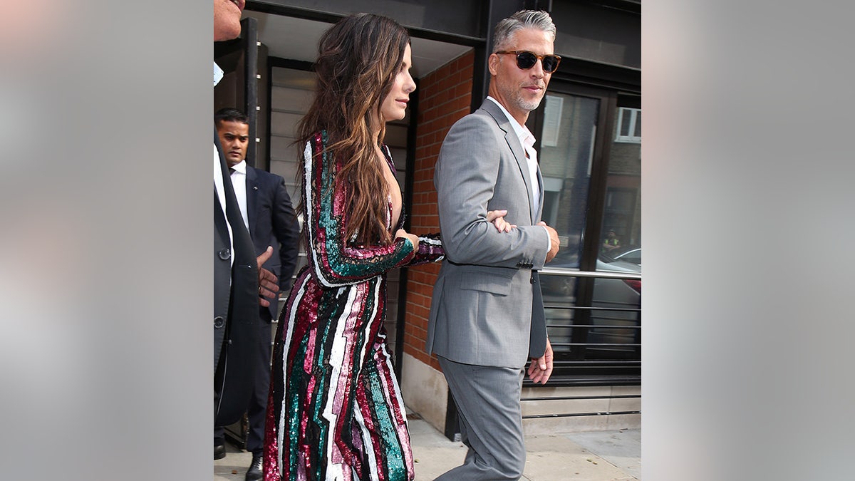 Sandra Bullock in multi-colored striped dress holds onto the arm of Bryan Randall in a grey suit