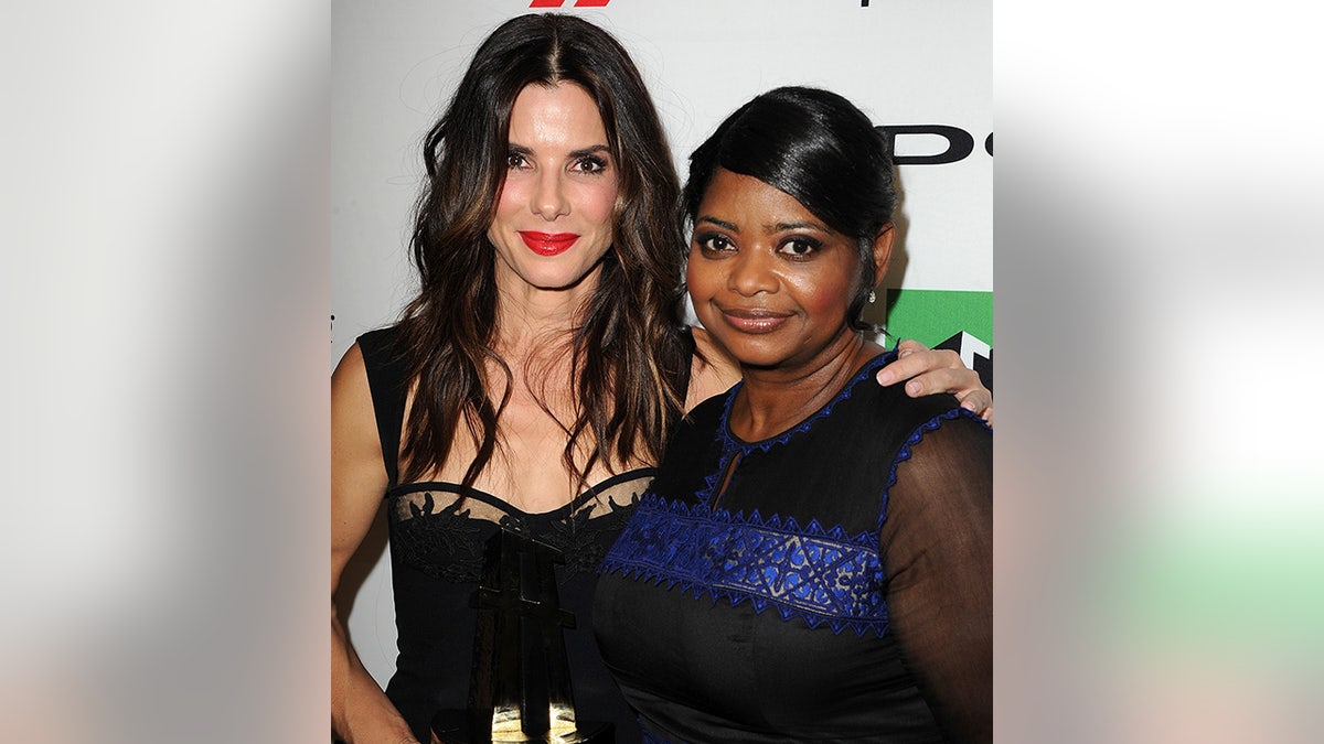 Sandra Bullock in a black dress puts her arm around Octavia Spencer in a dark dress with a blue stripe