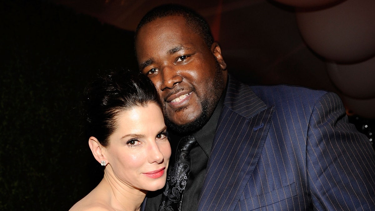 Sandra Bullock leans against Quinton Aaron in a blue pinstripe suit and black shirt and tie