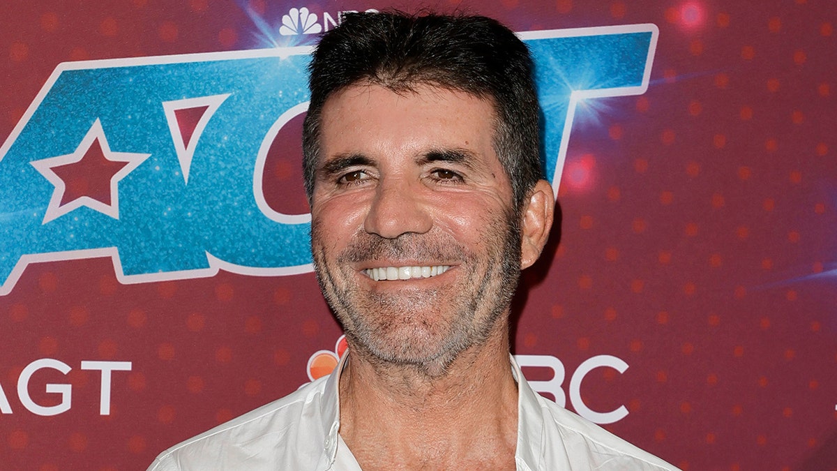 Simon Cowell smiles slightly to his right in a white shirt on the 'AGT' carpet