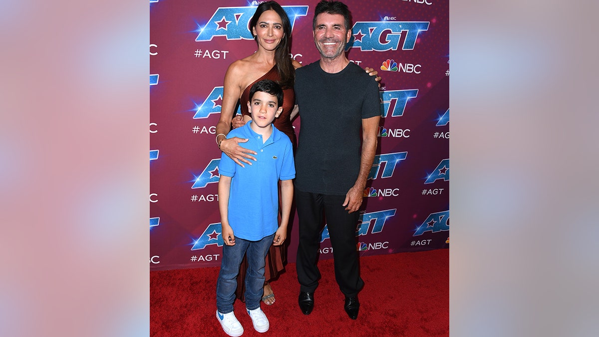 Simon Cowell in a black shirt and pants smiles with his partner Lauren Silverman and their son Eric in a blue polo shirt and jeans on the"AGT" carpet
