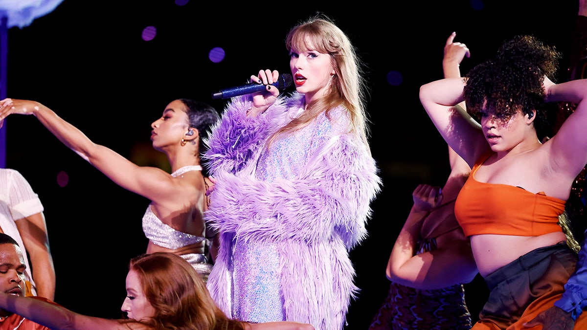 Taylor Swift sings on stage during "The Eras Tour" in a purple sparkle dress and feather lavender jacket