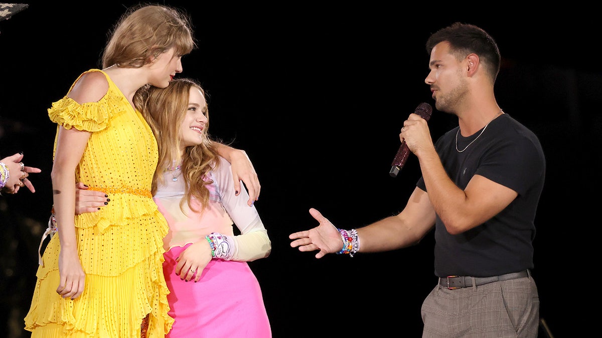 Taylor Swift in a bright yellow dresses hugs Joey King on stage while Taylor Lautner in a black shirt talks into the microphone during The Eras Tour in Missouri