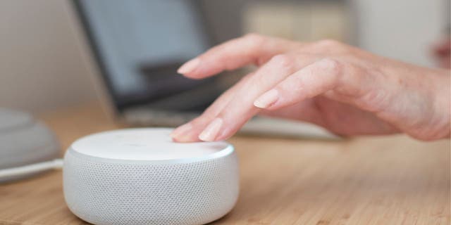 Açık renkli ahşap bir masanın üzerinde beyaz Amazon Alexa cihazı ve cihaza dokunan bir el