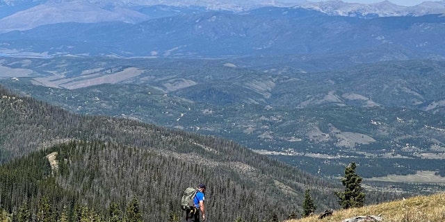A man and a dog search on a trail with mountains in the background