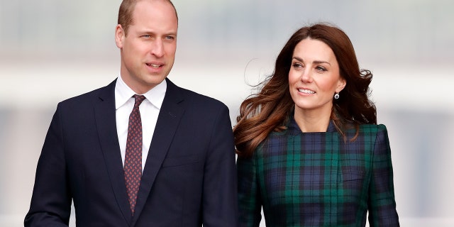 A close-up of Prince William wearing a dark blue blazer with a white shirt and red tie standing next to Kate Middleton in a green plaid dress
