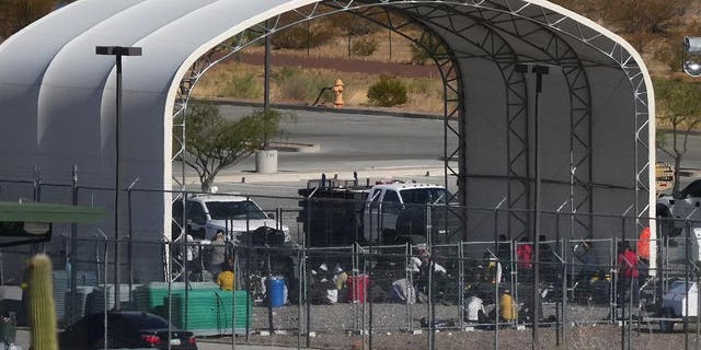 Migrants sit on the ground behind the fence under a fabric arched canopy b