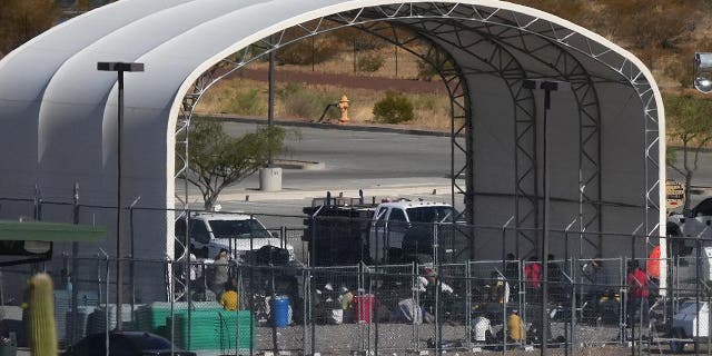 Migrants sit on the ground behind fencing under a fabric arched canopy b